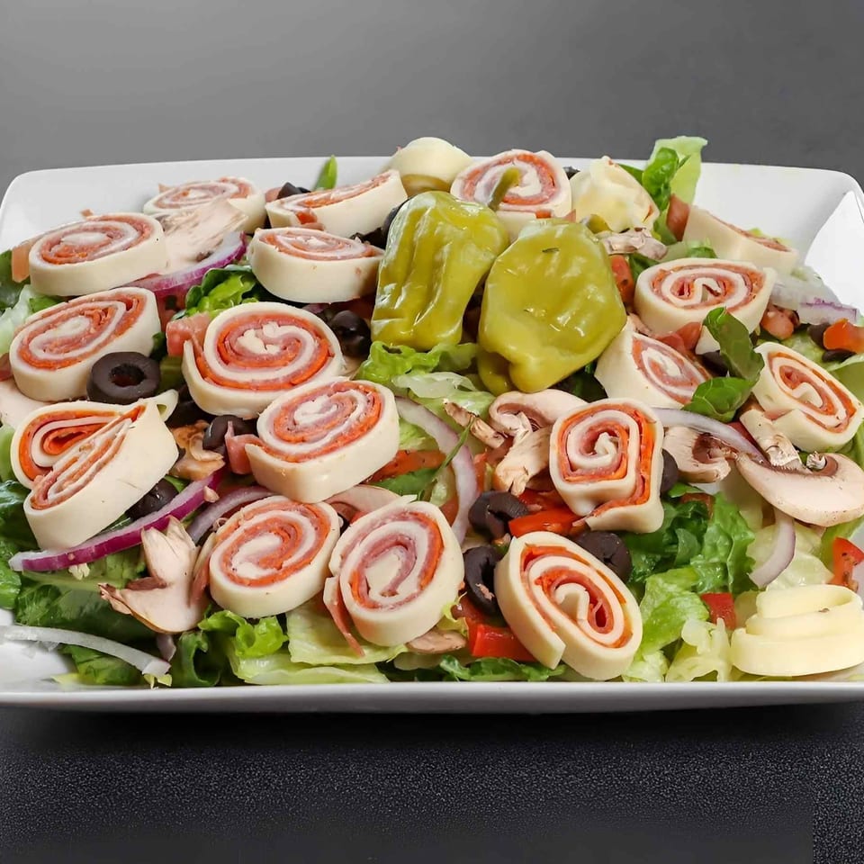 A white platter holds green salad topped with sliced Italian pinwheel wraps, banana peppers, mushrooms, olives, tomatoes, and onions.