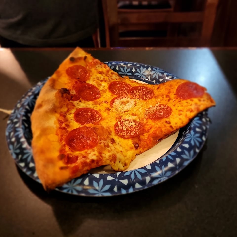Pepperoni pizza slice with melted cheese and grated cheese on a blue patterned plate at a restaurant table.
