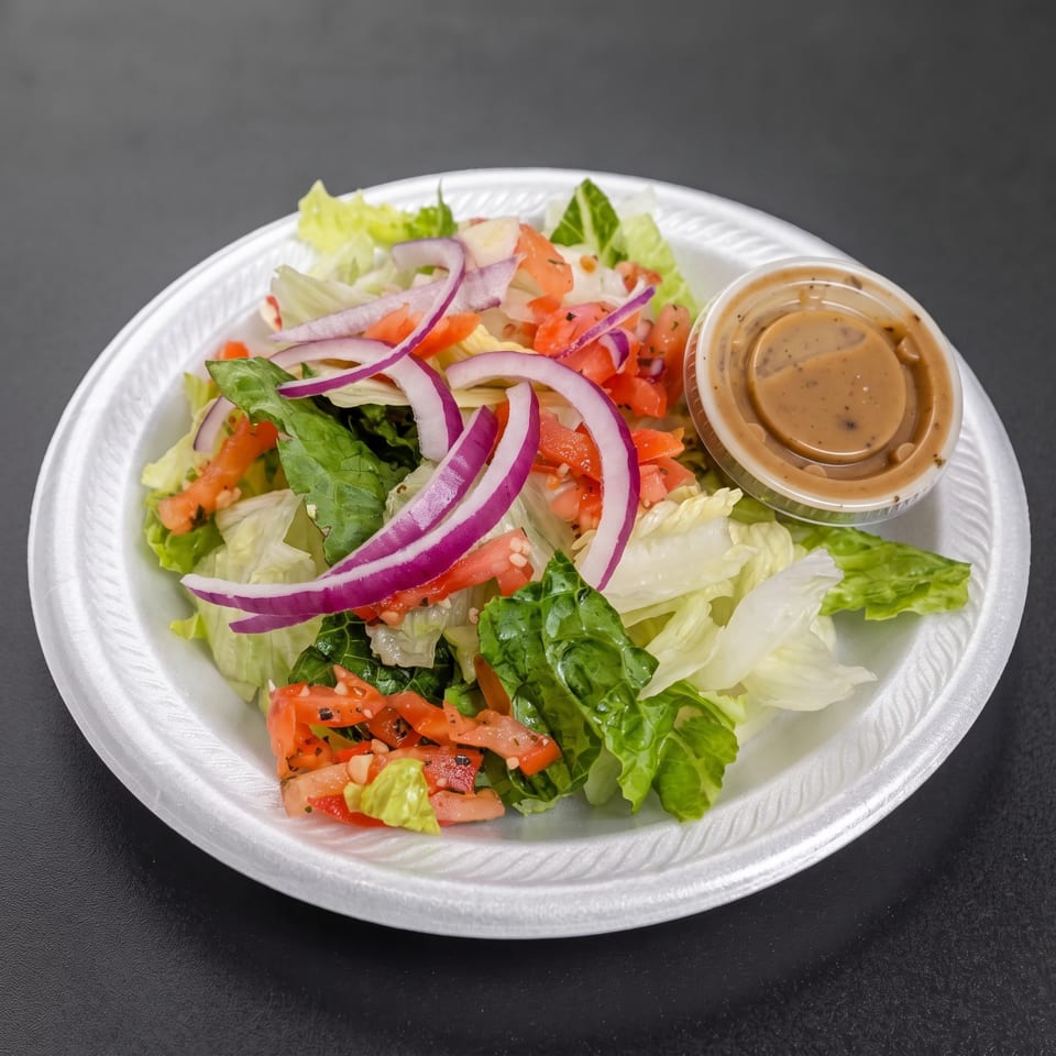 Fresh green salad with lettuce, tomatoes, red onions, and a cup of creamy dressing on a white plate.