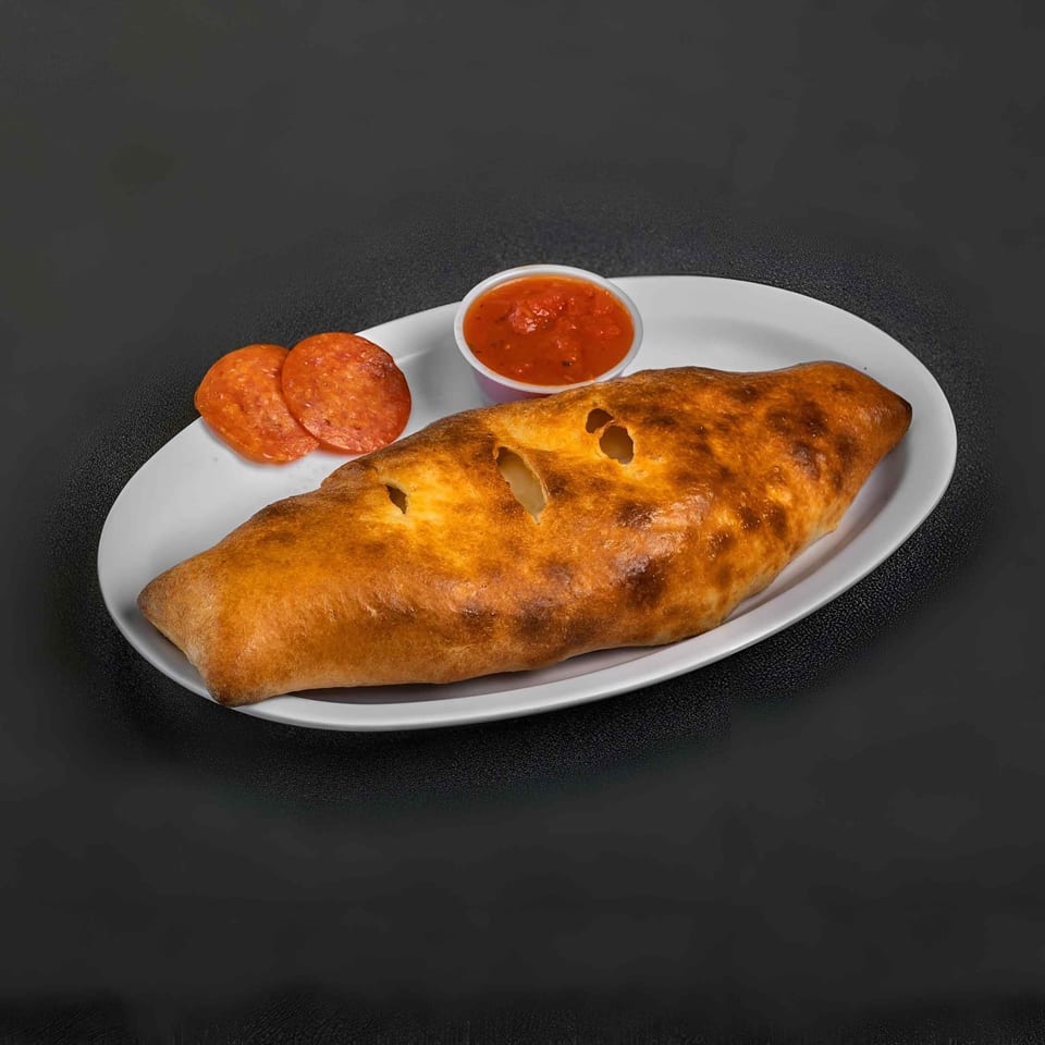 Golden-brown calzone served with sliced pepperoni and a cup of red dipping sauce on a white plate.