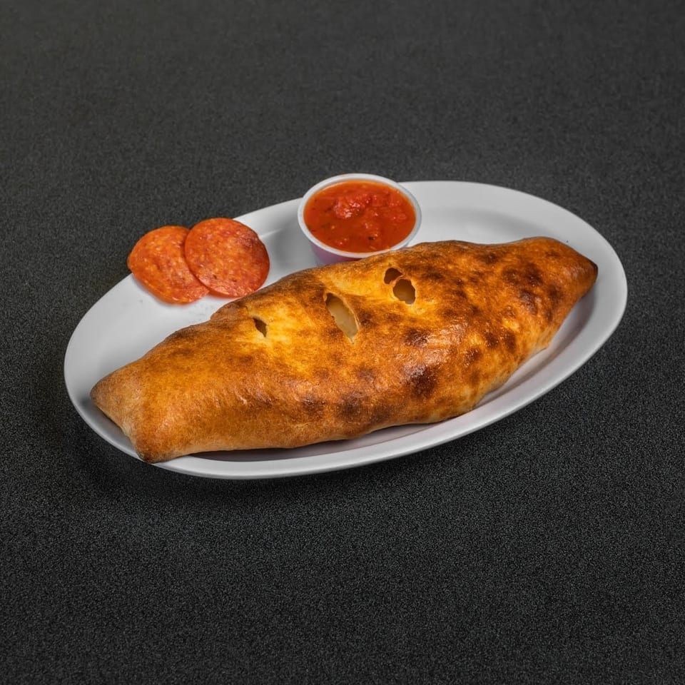 Golden-brown calzone served on a white plate with sliced pepperoni and a cup of marinara sauce.